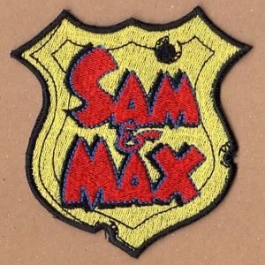 Può includere: Una toppa ricamata gialla e nera con le parole "Sam & Max" in lettere rosse con contorni blu.