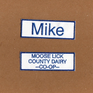 May include: Two rectangular embroidered patches with white fabric and blue borders. The top patch features the name Mike in blue embroidery. The bottom patch displays the text MOOSE LICK COUNTY DAIRY --CO-OP-- in blue embroidery.