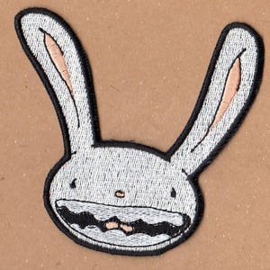 Op de afbeelding: Witte geborduurde patch van een cartoonkonijn met een brede grijns en grote oren. Het konijn heeft een zwarte omtrek en een zwarte mond.