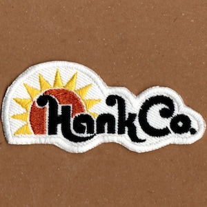 May include: Embroidered patch with the text "Hank Co." in a black, stylized font. The patch features a sun design with yellow rays and a red and orange center, all outlined in white.