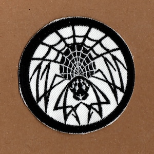 以下が含まれることがあります： クモの巣の中にクモが描かれた、白黒の刺繍パッチ。クモは正面を向いており、8本の脚があります。クモの巣は円形で、黒い輪郭があります。
