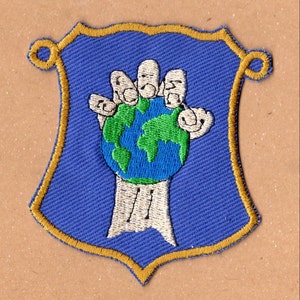 Puede incluir: Un parche bordado azul con un borde dorado en forma de escudo. El parche presenta una mano blanca sosteniendo un globo terráqueo verde y azul.
