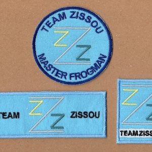 Puede incluir: Tres parches de tela azul con el texto "Team Zissou" y un diseño estilizado en "Z". El parche superior es redondo y dice "Master Frogman". El parche del medio es rectangular y dice "Team Zissou". El parche inferior es cuadrado y dice "Team Zissou".