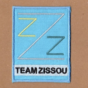 Könnte beinhalten: Ein blauer Stoff-Patch mit einem stilisierten Buchstaben Z in gelber, grauer und grüner Farbe. Der Text "TEAM ZISSOOU" ist in Schwarz unter dem Buchstaben gedruckt.