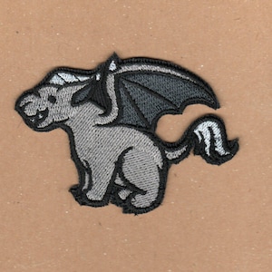 Könnte beinhalten: Ein gestickter Aufnäher mit einer Cartoon-Kreatur mit Flügeln und einem Horn. Die Kreatur ist grau mit schwarzem Umriss und einem weißen Horn.