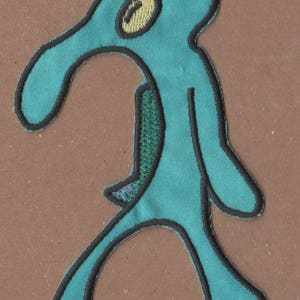 May include: A blue and black embroidered patch of a cartoon character with a long nose and one eye. The character is walking with its arms outstretched.