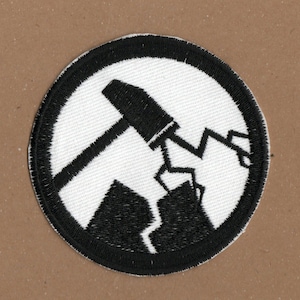 May include: Black and white embroidered patch with a circular design. The centre of the circle features a black silhouette of a hammer breaking a rock.