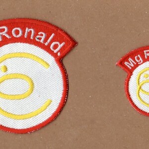 May include: Two embroidered patches with a white circle and yellow letter C inside. The larger patch has a red curved banner with the text "Mg Ronald." The smaller patch has a red curved banner with the text "Mg Ronald."
