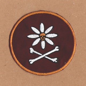 May include: A round, maroon patch with an orange border. The patch features a white daisy with a yellow centre and a skull inside the centre. Two white crossed bones are below the daisy.