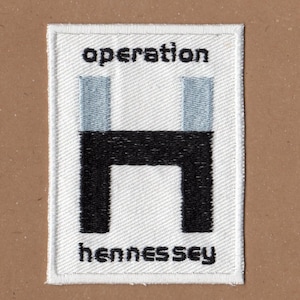 Puede incluir: Parche blanco bordado con texto negro que dice "operation hennesse" y una letra "H" estilizada en negro y gris en el centro.
