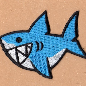 May include: A blue embroidered shark with white teeth and a black outline. The shark is smiling with its mouth open.