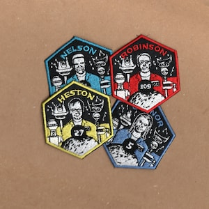 May include: Four hexagonal embroidered patches with black outlines and white stitching. Each patch features a different person with a number and a name. The names are "Nelson", "Robinson", "Heston", and "Nor".