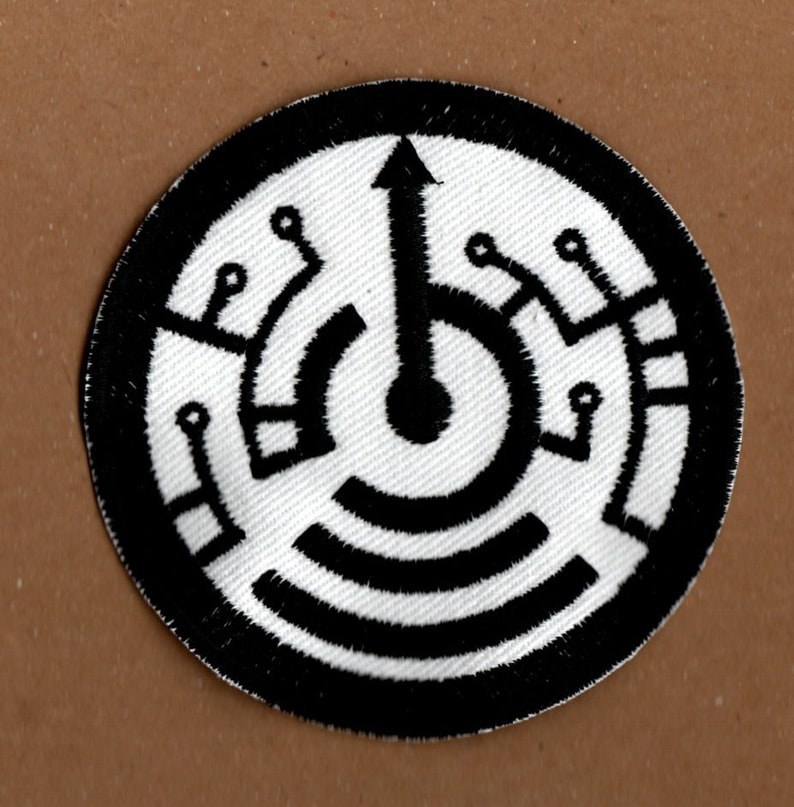 May include: Black and white embroidered patch with a circular design. The centre of the circle features a black arrow pointing upwards. The arrow is surrounded by a series of concentric circles and lines.