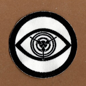 May include: Black and white embroidered patch featuring a stylised eye design. The eye has a target-like pattern inside with three arrows pointing towards the centre.