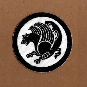 Könnte beinhalten: Schwarz-weiß gestickter Aufnäher mit einer stilisierten Fabelwesen mit Flügeln, das einem Greif ähnelt, auf einem weißen runden Hintergrund mit schwarzem Rand.