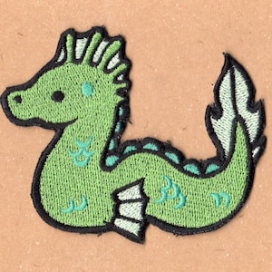 以下が含まれることがあります： 白のたてがみと尾びれを持つ緑色の刺繍されたタツノオトシゴのパッチ。タツノオトシゴは黒い輪郭と、濃い緑色の模様のある薄い緑色の体を持っています。