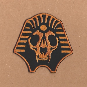 May include: An embroidered patch featuring a black cat skull with an orange outline, inside a gold pharaoh's headdress.
