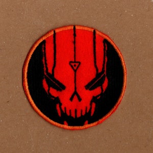 Puede incluir: Un parche bordado rojo y negro con un diseño de calavera. La calavera tiene un símbolo triangular en el centro y tres líneas verticales por encima.