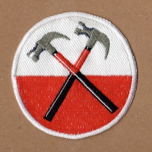 Puede incluir: Un parche circular blanco y rojo con dos martillos cruzados en gris y negro. Los martillos están en el centro del parche y están cruzados sobre un fondo blanco. La mitad inferior del parche es roja.