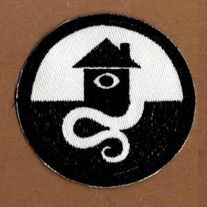 May include: Black and white embroidered patch featuring a house with an eye and a snake design.