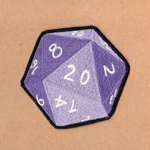 Peut inclure: Dé à 20 faces de couleur violette avec des chiffres blancs, brodé sur un patch en tissu.