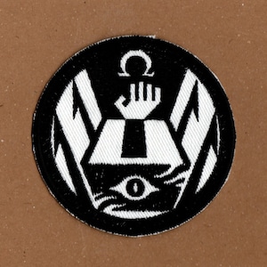 May include: Black and white embroidered patch featuring a stylised eye, hand, and omega symbol. The patch is circular and has a black background with white details.
