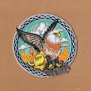 May include: Embroidered patch featuring a colorful griffin with a lion body and eagle wings, set against a blue and green background with clouds and a Celtic knot border.