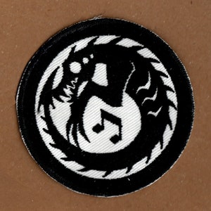 May include: Black and white embroidered patch featuring a stylised monster with musical notes. The monster is depicted in a circular design with a black outline.