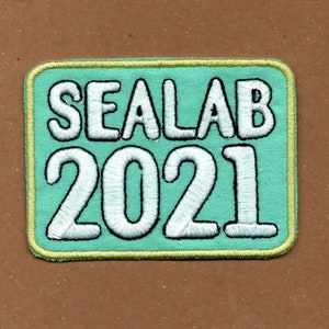 May include: A teal blue embroidered patch with a yellow border. The patch says "SEALAB 2021" in white lettering.