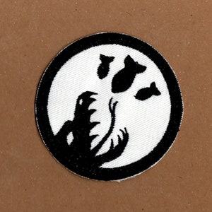 May include: Black and white embroidered patch with a circular design. The centre of the circle features a silhouette of a monster with its mouth open, and three bombs flying above it.