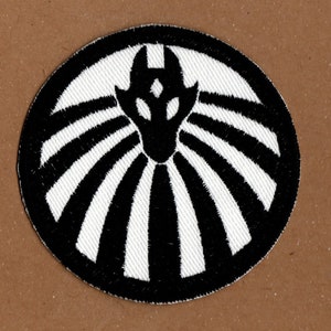 Op de afbeelding: Zwart-witte geborduurde patch met een gestileerd wezen met een enkel oog en een stralend patroon.