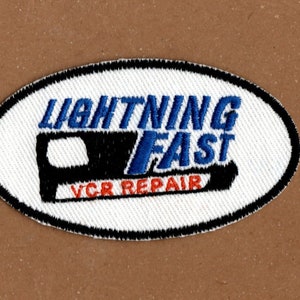 May include: White oval patch with black and blue embroidery. The text "Lightning Fast VCR Repair" is embroidered on the patch.
