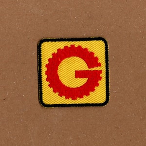 May include: A yellow and black embroidered patch with a red gear-shaped letter "G" in the center.
