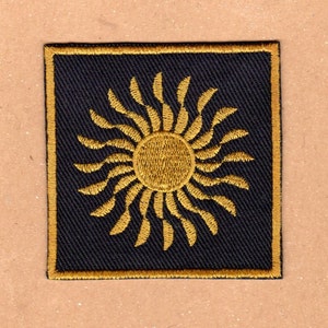 Op de afbeelding: Een zwarte geborduurde patch met een gouden zonontwerp. De zon heeft een cirkelvormig midden en stralende stralen.
