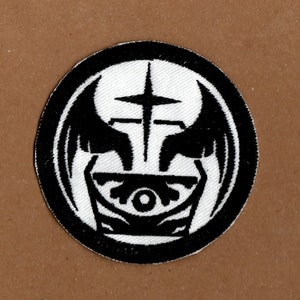 May include: Black and white embroidered patch featuring a stylised eye, wings, and cross design. The eye is surrounded by a circle with a cross above it. The wings are on either side of the eye and cross.