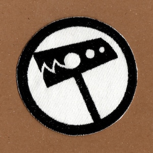 May include: A white embroidered patch with a black circle around the edge. Inside the circle is a black silhouette of a hammer with a jagged edge and three circles on the top.