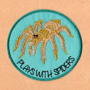 May include: A round, turquoise blue patch with a yellow and brown embroidered spider in the center. The text "Plays with Spiders" is embroidered in black around the spider.