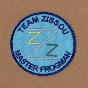 Può includere: Una toppa circolare blu e bianca con il testo "Team Zissou Master Frogman" che circonda una lettera "Z" stilizzata in giallo, grigio e verde.