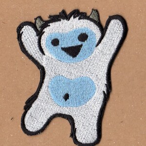 Op de afbeelding: Witte geborduurde patch van een glimlachende yeti met blauwe accenten. De yeti staat met zijn armen omhoog.