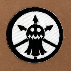 May include: Black and white embroidered patch featuring a ghost-like figure with a toothy grin and four arrows pointing outwards. The patch is circular and has a black border.