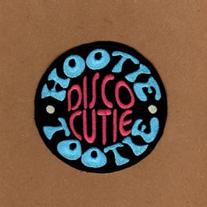 Puede incluir: Un parche redondo y negro bordado con las palabras "Disco Cutie Booty" en rosa y azul claro. El parche tiene un diseño retro y juguetón, perfecto para añadir a ropa o accesorios.
