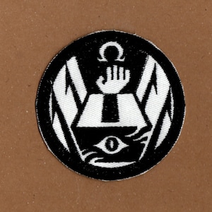 May include: Black and white embroidered patch featuring a stylised eye, a hand reaching upwards, and an omega symbol. The design is enclosed by two wings.