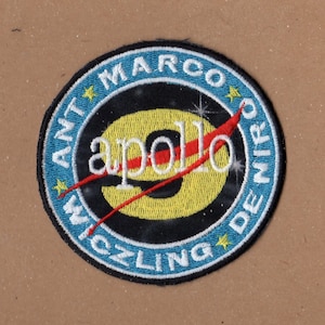 May include: A blue and white embroidered patch with a yellow and red "Apollo 9" logo. The text around the logo reads "Wichling De Marco" and has stars.
