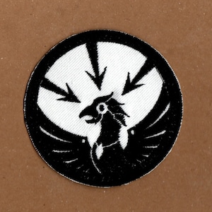 May include: Black and white embroidered patch featuring a stylised bird with wings spread, surrounded by a circle of arrows pointing inwards.