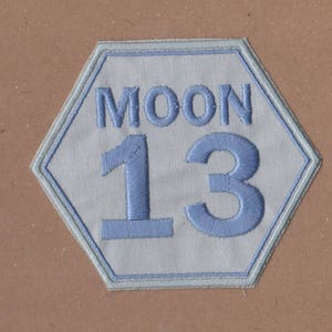 May include: A white hexagonal patch with a blue embroidered border. The patch has the word "MOON" and the number "13" in blue embroidery.