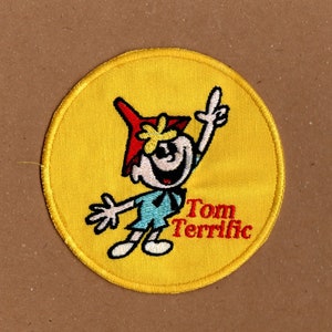 Könnte beinhalten: Gelber gestickter Aufnäher mit einer Comicfigur, die einen roten und gelben Hut trägt. Die Figur lächelt und hebt einen Arm. Der Text "Tom Terrific" befindet sich unter der Figur.
