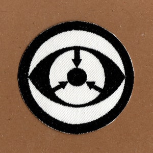 May include: Black and white embroidered patch featuring an eye design. The eye has three arrows pointing inwards towards a black circle in the centre.