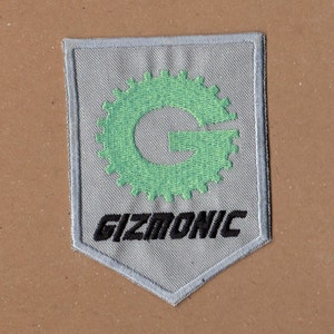 May include: A gray embroidered patch with a green gear-shaped logo featuring the letter "G" and the text "GIZMONIC" in black.
