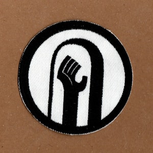 May include: Black and white embroidered patch with a raised fist inside a circle. The fist is raised in the air with the thumb pointing towards the viewer.
