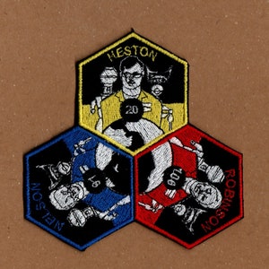 May include: Three hexagonal embroidered patches with black backgrounds and white outlines. The top patch is yellow with the text "HESTON" and a cartoon image of a man with glasses. The left patch is blue with the text "NELSON" and a cartoon image of a person. The right patch is red with the text "ROBINSON" and a cartoon image of a person.